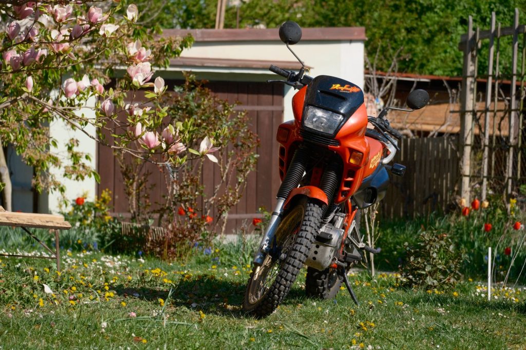 Honda NXR160 Bros 2027 chega com novas cores e reforço em itens de segurança Motocicleta laranja estacionada em um jardim florido.