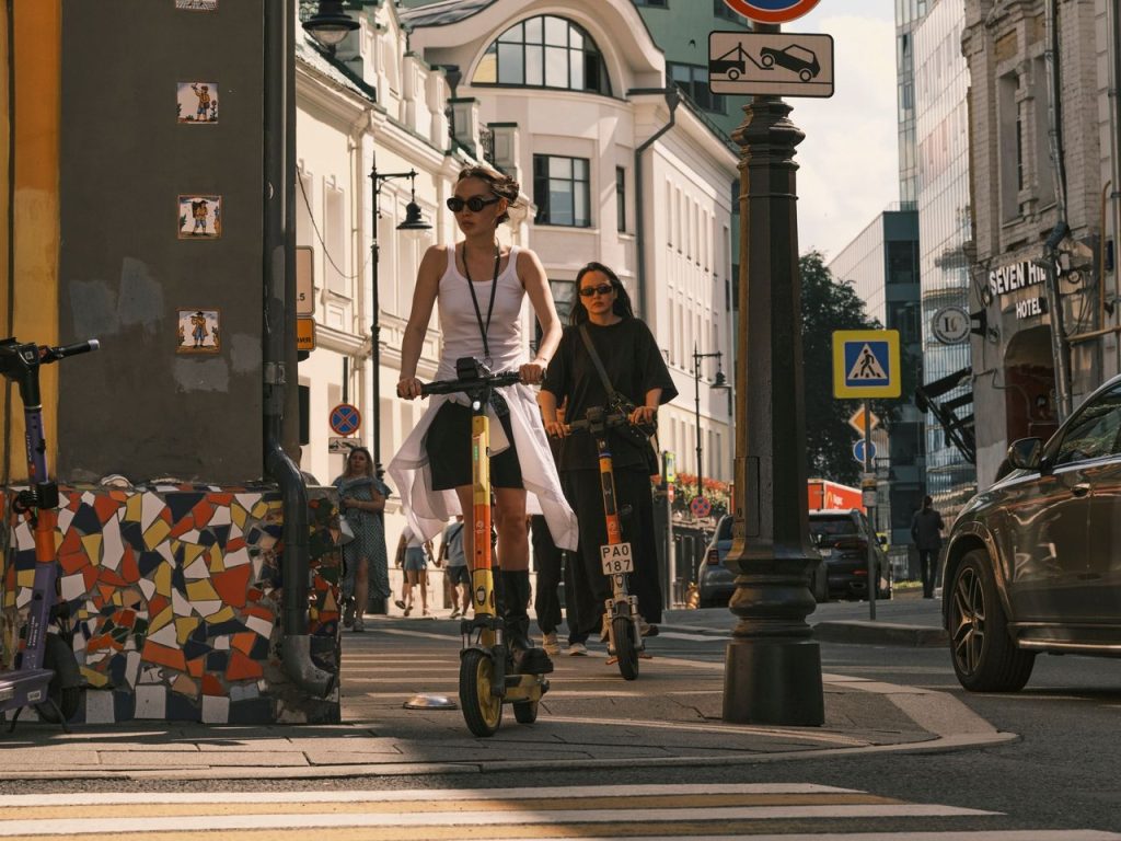 Duas mulheres conduzem scooters elétricas em uma rua urbana durante um deslocamento.