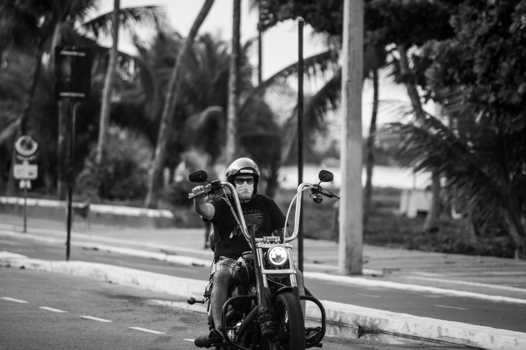 Motociclista usando capacete e máscara circula de moto por uma rua urbana no Brasil.